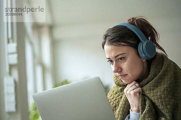 Woman with wireless headphones wrapped in blanket looking at laptop