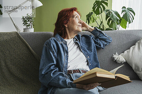 Thoughtful senior woman reading book sitting on sofa at home