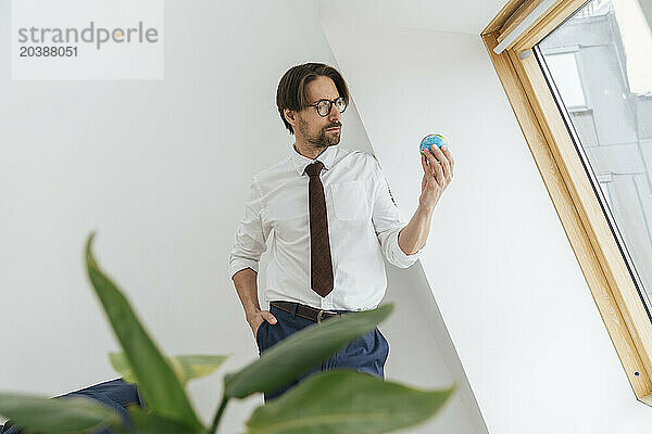 Confident businessman holding globe leaning on white wall at office