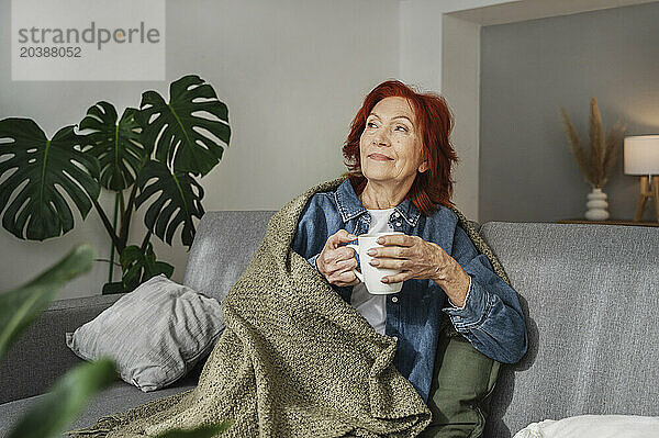 Senior woman sitting relaxed under blanket on sofa drinking tea at home