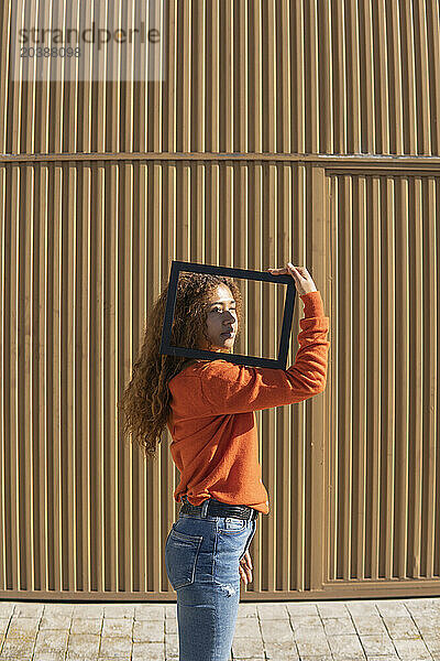 Young woman holding picture frame on shoulder standing in front of metal wall