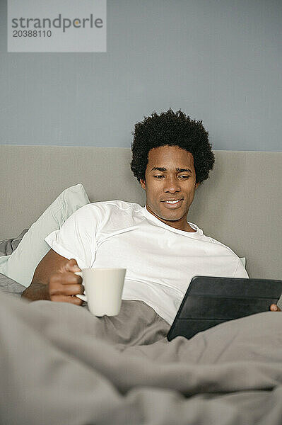 Smiling man with coffee cup using tablet PC relaxing on bed in bedroom at home