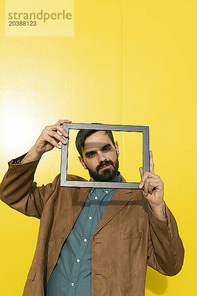 Smiling man wearing brown jacket and looking through photo frame