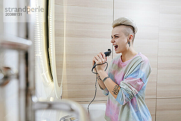 Young woman singing in bathroom holding electric razor at home