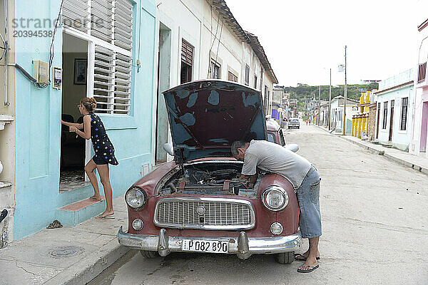 Cuba  Gibara une femme rentre dans sa maison alors qu’un homme répare le moteur de sa vieille voiture américaine des années 50 / Cuba  Gibara  a woman enters her house while a man is fixing the engine of his old 50s American car