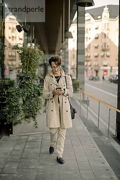 Full length of retired senior woman using smart phone while walking on sidewalk