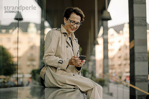 Smiling retired senior woman using smart phone while sitting on bench