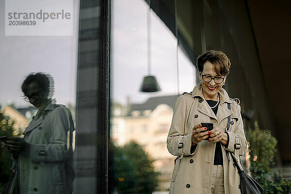 Smiling senior woman using smart phone while standing by glass reflection