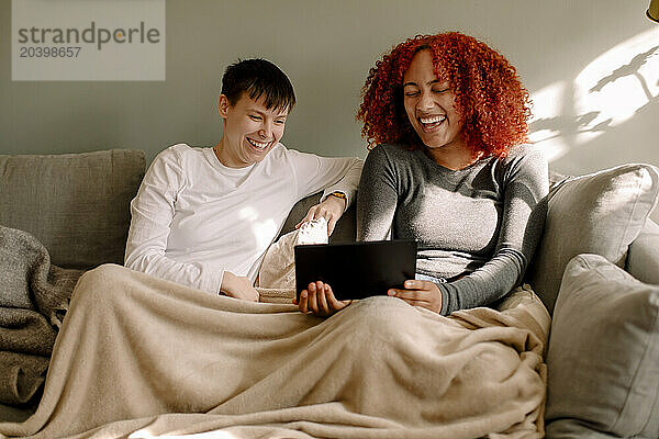 Cheerful young women sharing laptop while watching movie together sitting on sofa at home