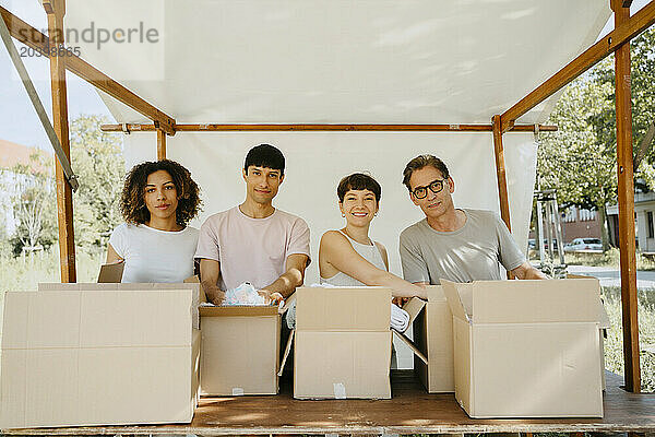 Portrait of smiling male and female volunteers standing under tent with cardboard boxes on table at community center