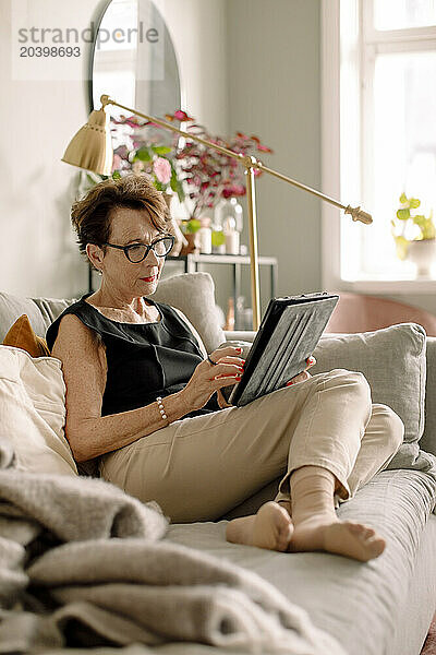 Full length of retired senior woman using digital tablet while sitting on sofa at home