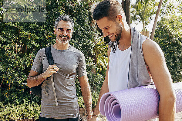 Portrait of smiling man standing with male friend holding yoga mat at wellness resort