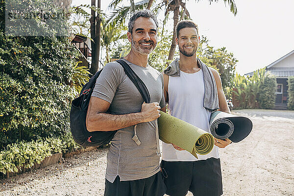 Smiling man carrying backpack while standing with male friend holding yoga mat at wellness resort