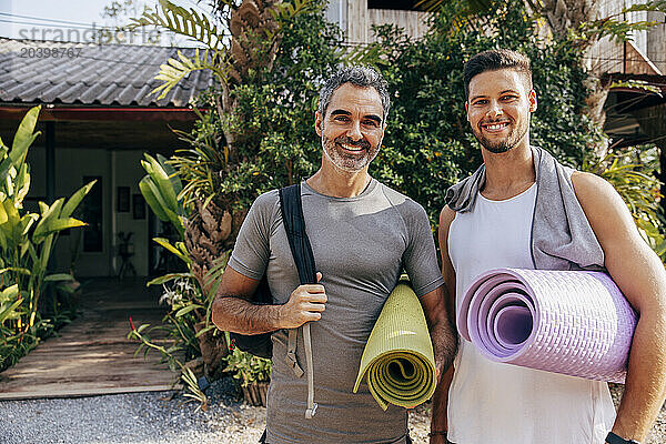 Portrait of smiling men holding yoga mats while standing in front of wellness resort