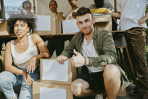 Portrait of smiling male volunteer showing thumbs up gesture while crouching by female colleague near boxes at community