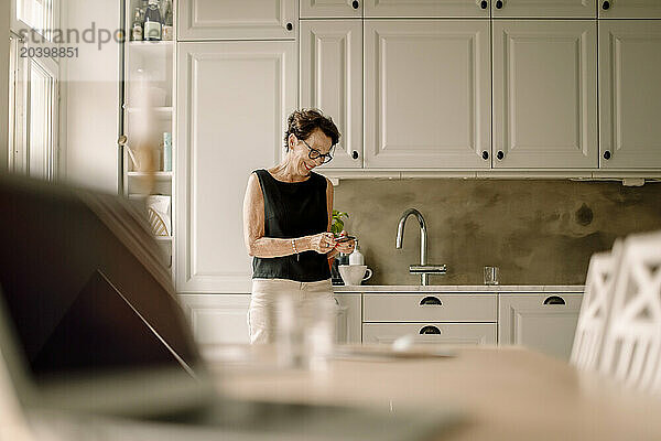 Retired senior woman using smart phone while leaning on kitchen counter