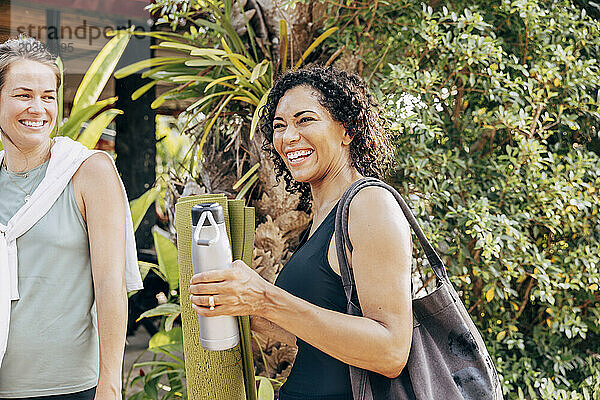 Happy woman holding water bottle while standing near female friend at wellness resort