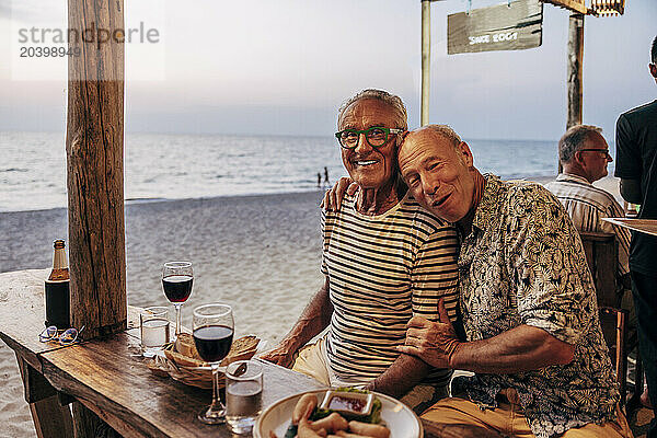 Portrait of happy gay couple with wine and snacks on table at beach restaurant