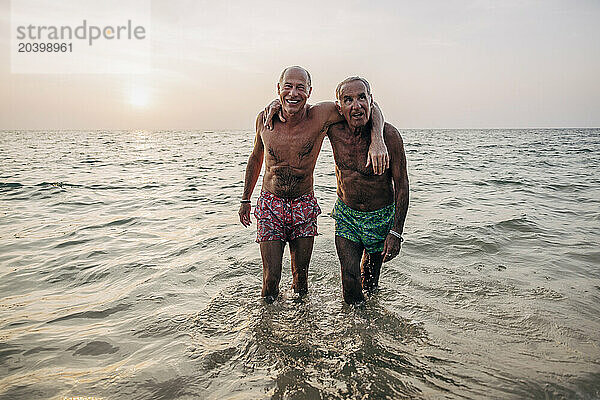 Portrait of shirtless gay couple standing in sea on vacation at sunset