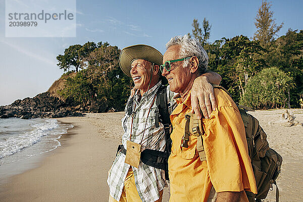 Happy senior gay couple standing with arms around and looking at sea on beach