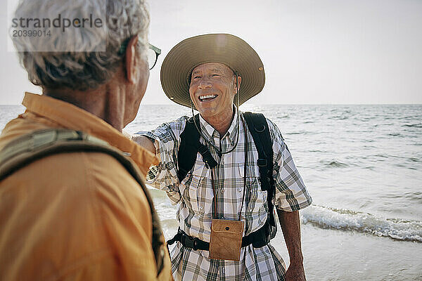 Happy senior gay man talking with boyfriend while standing near sea at beach on vacation