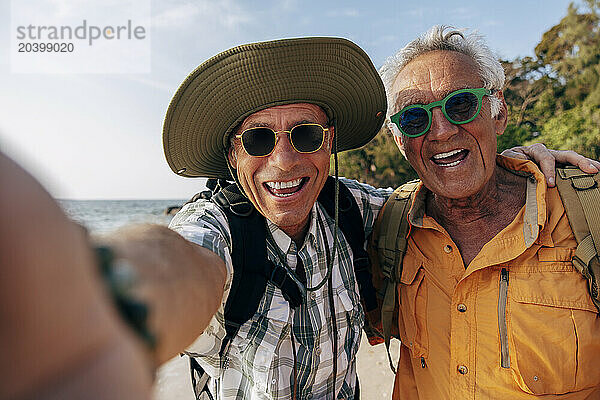 Portrait of happy senior gay couple wearing sunglasses and taking selfie at beach