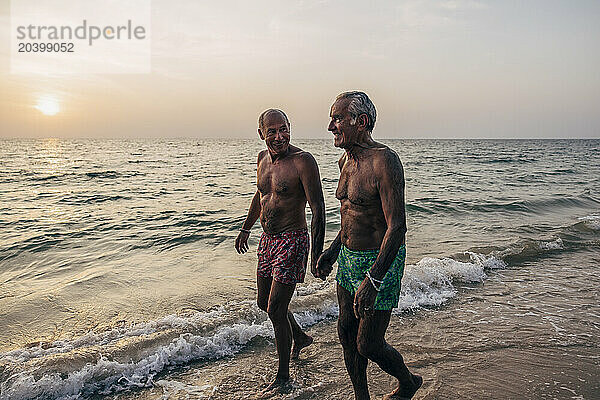 Shirtless gay couple holding hands while walking on shore at beach