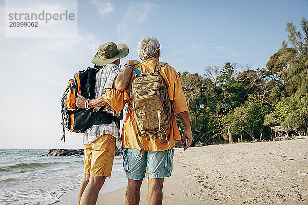 Low angle rear view of senior gay couple with backpacks standing at beach on vacation