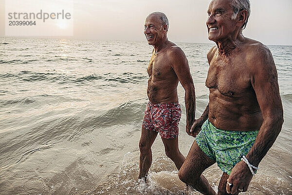 Shirtless senior gay couple holding hands while walking in sea at beach