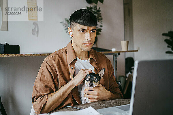 Serious male freelancer stroking dog while looking at laptop in home office