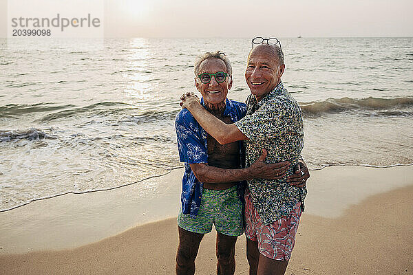 Portrait of smiling senior gay couple embracing each other while standing near shore at beach