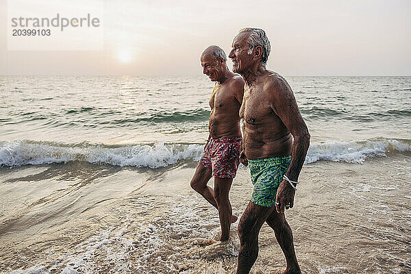 Senior shirtless gay couple holding hands while walking near shore at beach against sky