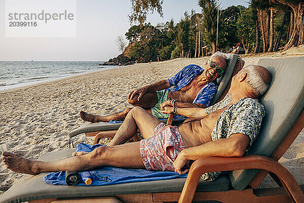 Happy senior gay couple reclining on deck chair at beach during vacation