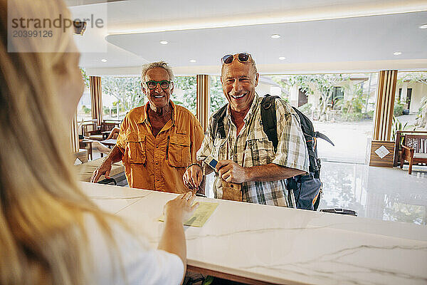 Happy senior gay couple talking with female receptionist at desk in hotel