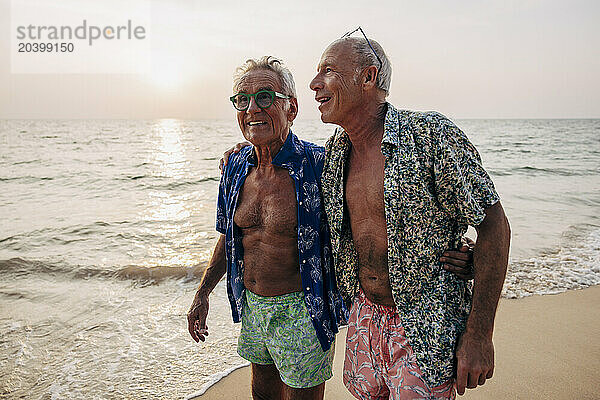 Carefree senior gay couple walking with arms around near shore at beach