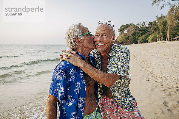 Senior gay man kissing boyfriend on vacation at beach