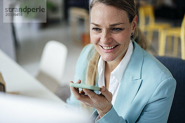 Smiling mature businesswoman talking through smart phone speaker at office