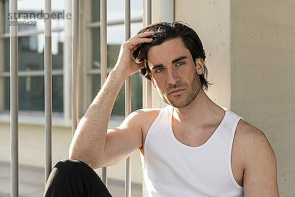 Handsome young man wearing undershirt sitting with hand in hair by fence