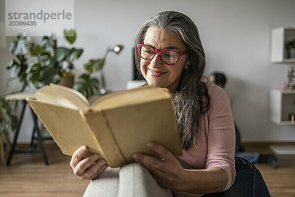 Smiling mature woman reading book sitting on sofa in living room at home