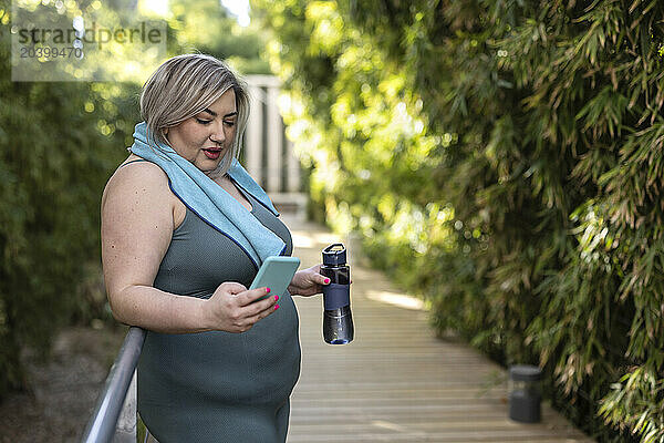 Curvy young woman standing with water bottle and using smart phone near plants