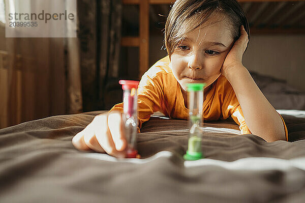 Thoughtful cute boy looking at hourglass on bed at home