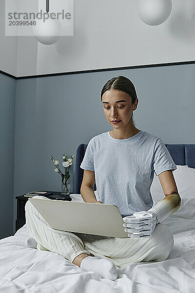 Woman with arm prosthesis using laptop sitting on bed in bedroom at home