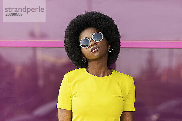 Confident young Afro woman wearing sunglasses in front of magenta window