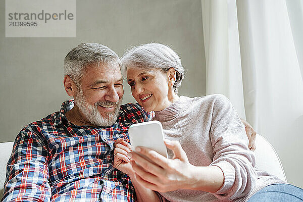 Smiling mature couple with smart phone sitting on couch at home