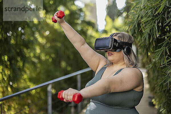 Young curvy woman wearing virtual reality simulator and exercising with dumbbells near plants in park