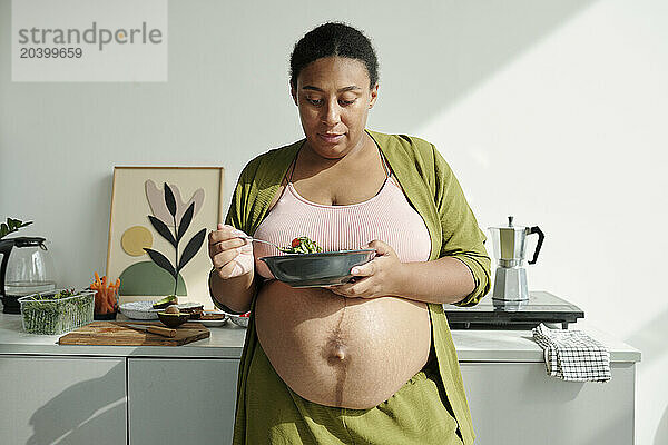 Plus size pregnant woman eating salad with fresh vegetables in kitchen at home