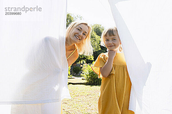 Smiling mother and daughter behind white bedsheet hanging on clothesline at back yard