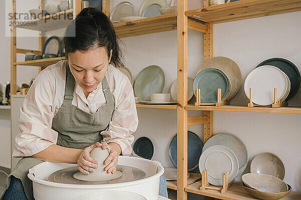 Confident craftsperson molding clay on pottery wheel by rack at art studio