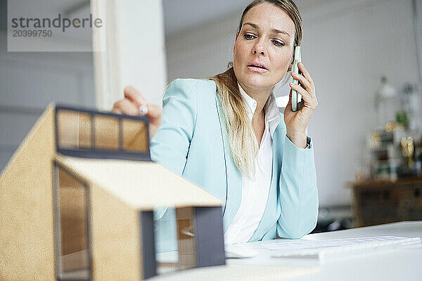 Confident mature architect talking on mobile phone sitting with model house at desk in office