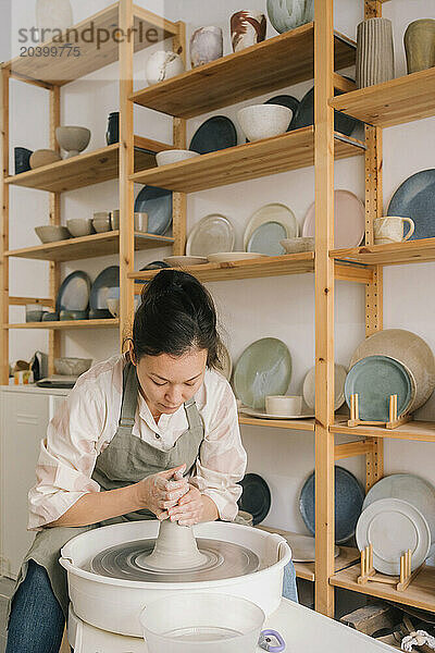 Confident craftsperson molding clay on pottery wheel by rack at workshop